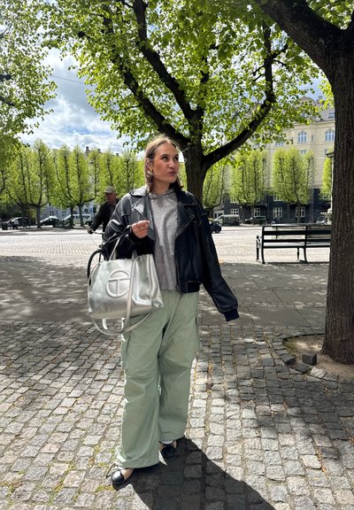 Chaqueta de cuero negra, suéter gris, pantalones cargo verde claro. Bolso plateado con logotipo y bailarinas negras. Fondo de pavimento de adoquines y árboles.