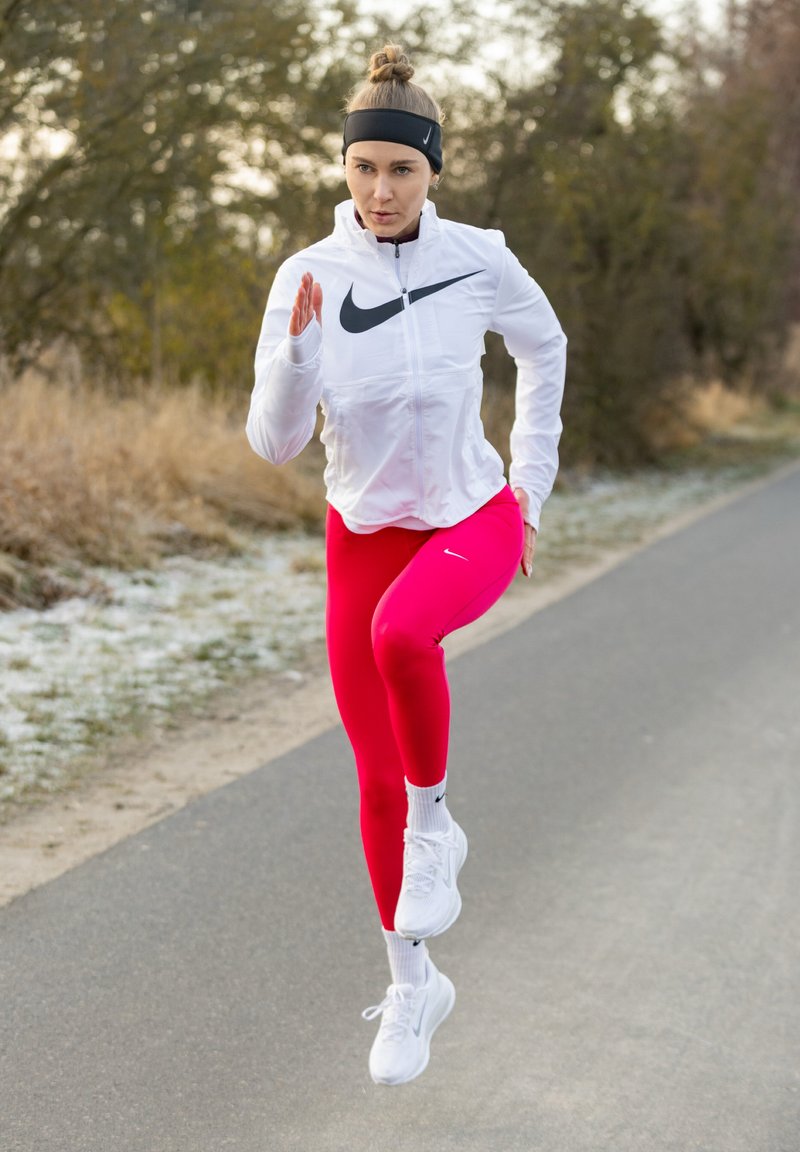 Giacca bianca con cerniera e logo Nike nero, leggings rosa acceso e scarpe da ginnastica bianche. Il corridore si muove con energia su un sentiero.