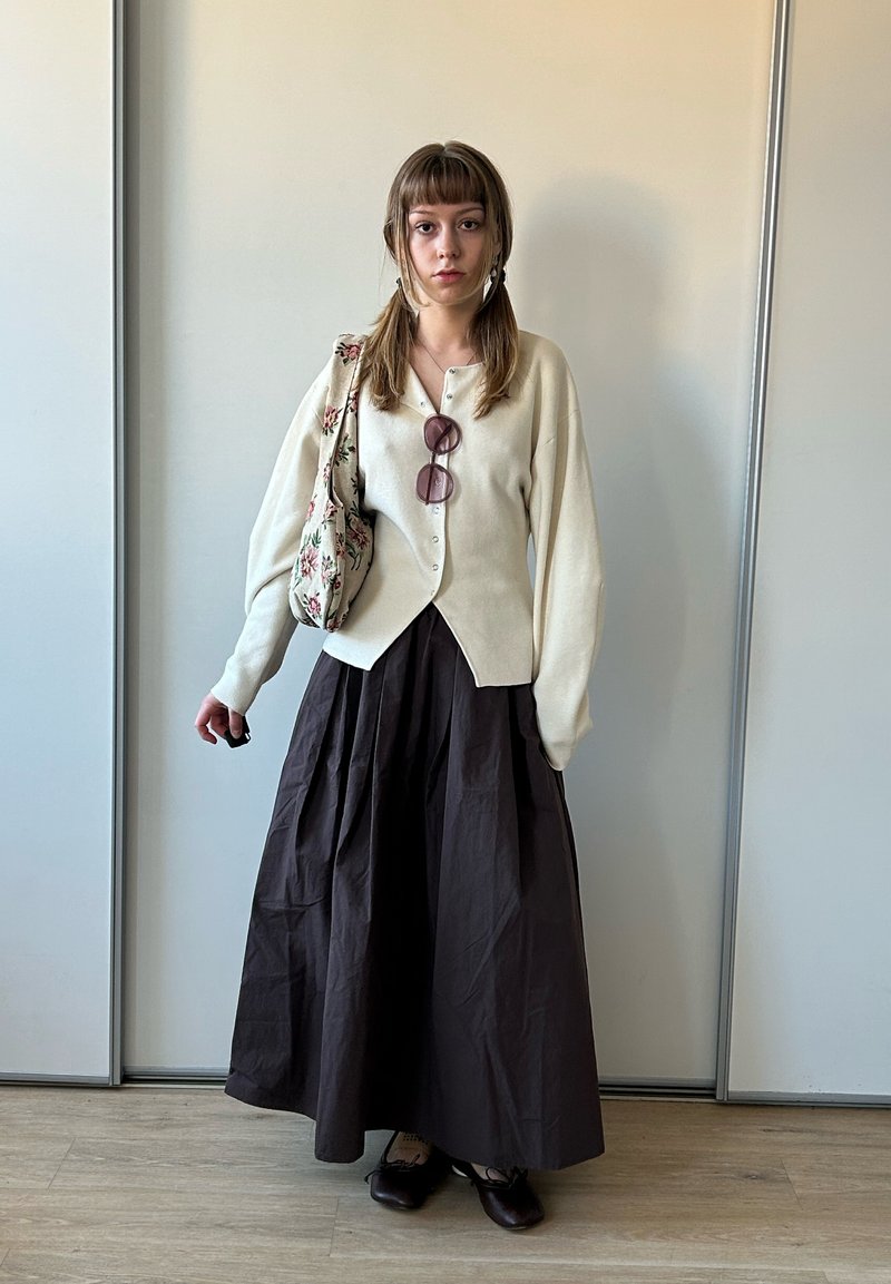 Jeune femme aux longs cheveux en couettes porte un cardigan crème, une jupe plissée foncée, des chaussures noires, un sac fleuri à l'épaule et des lunettes de soleil roses sur la poitrine.