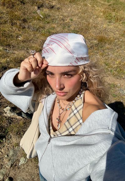 Pañuelo blanco con rayas diagonales rojas, sudadera gris con cremallera, blusa de cuadros beige, collares en capas, con cabello rubio rizado en un entorno natural.