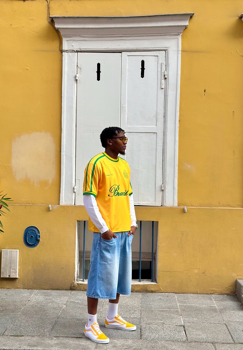 Uomo con maglietta sportiva gialla e pantaloncini di jeans in piedi su pavimentazione in pietra davanti a un muro giallo con persiane bianche chiuse.