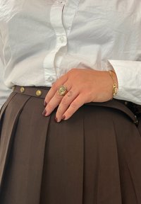 Brąspiająca spódnica z plisami z złotymi guzikami, zestawiona z białą koszulą z guzikami. Ręka ozdobiona złotym pierścionkiem i bransoletką, z tatuażami.