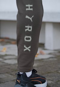 Pantalon de survêtement gris avec "HYROX" imprimé verticalement en couleur claire et effacée ; associé à des baskets noires avec des accents orange.