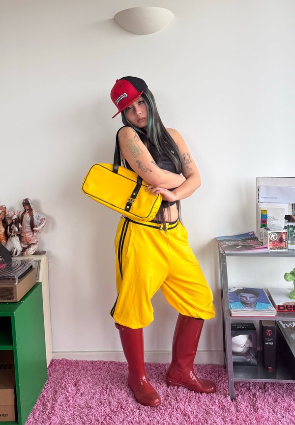 Jeune femme aux longs cheveux noirs portant une casquette rouge et noire, un pantalon jaune, des bottes rouges et portant un sac jaune contre un mur blanc.