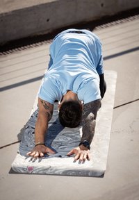 Mata do jogi z wirującym wzorem w odcieniach szarości i bieli. Osoba wykonująca skłon do przodu z wyprostowanymi ramionami, ubrana w jasnoniebieską koszulkę.