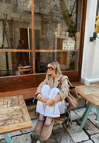 Femme avec des lunettes de soleil, blazer beige, pantalon blanc et chaussons assise sur une chaise en bois à l'extérieur d'un café près de tables et d'une vitre.