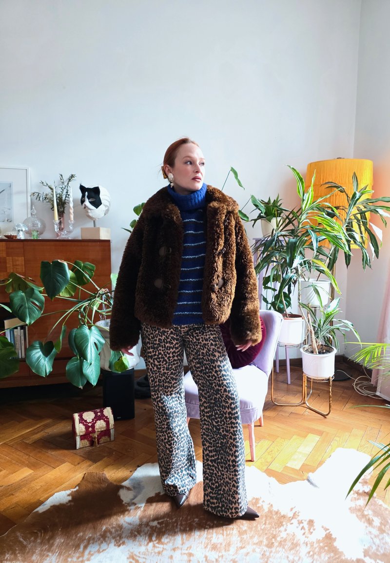 Femme en veste en fourrure marron et pantalon imprimé léopard se tient dans un salon lumineux avec des plantes, un fauteuil violet et un sol en bois.