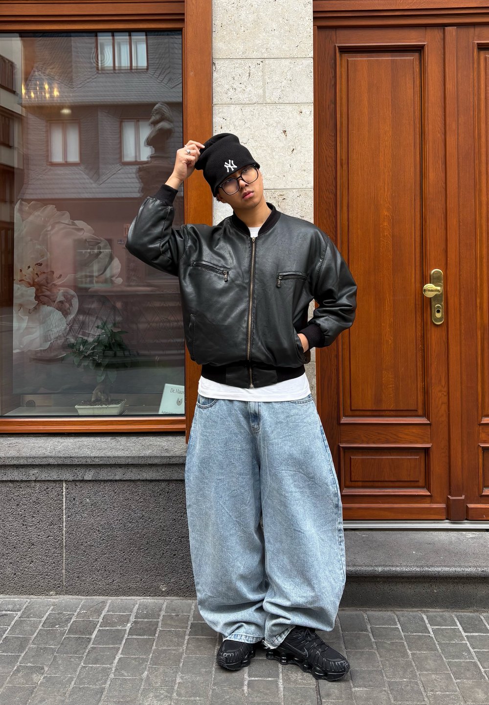 Jeune personne portant un bonnet noir, une veste en cuir noire, un jean bleu clair surdimensionné et des baskets noires, debout sur un trottoir en pierre près d'une porte en bois.
