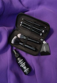 Un étui à pinceaux noir zippé contient plusieurs pinceaux de maquillage avec des poignées noires mates ; un spray de finition mate NYX est placé à proximité sur un tissu violet.