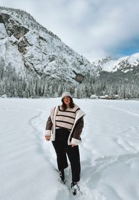 Una persona si trova in un terreno innevato indossando un maglione a righe spesso e una giacca imbottita marrone con fodera in shearling. Ha pantaloni neri e un cappello di maglia.