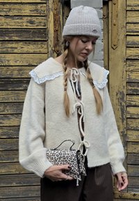 Krémový vlnený kardigan s dizajnom krajkového goliera, s prednými viazaniami. Drží malú kabelku s leopardím vzorom a rúčkou. Svetlo šedá pletená čiapka.
