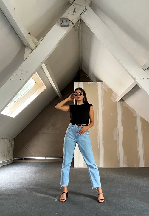 Egy fehér ujjatlan felsőt és menta zöld szoknyát viselő nő egy fonott székben ül, kezében egy pohárral, mellette egy üveg vízhűtő és cserepes növények vannak.