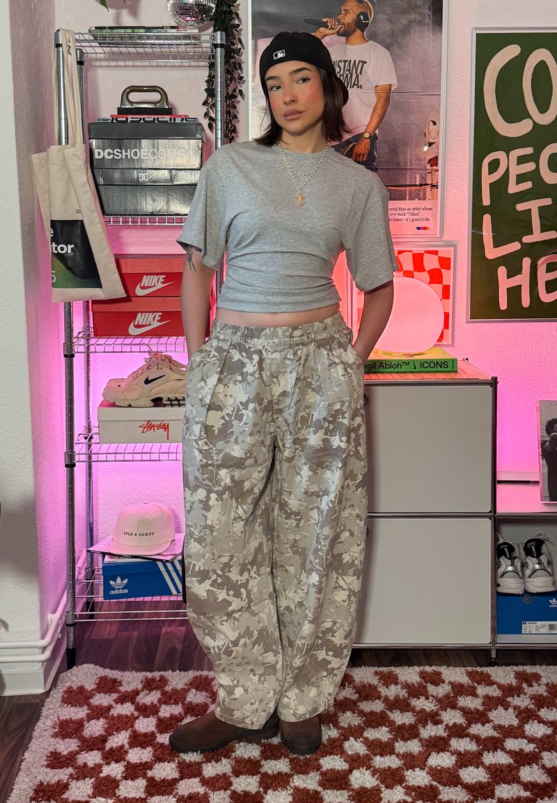 Jeune personne portant un haut gris court, une casquette noire et un pantalon camouflage beige debout dans une pièce avec des boîtes de baskets et des affiches.