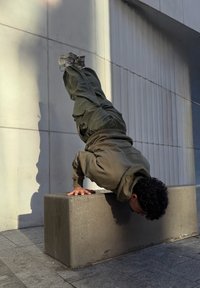 Une personne portant un sweat à capuche et un pantalon vert olive fait un poirier sur un bloc de pierre rectangulaire contre un mur texturé.