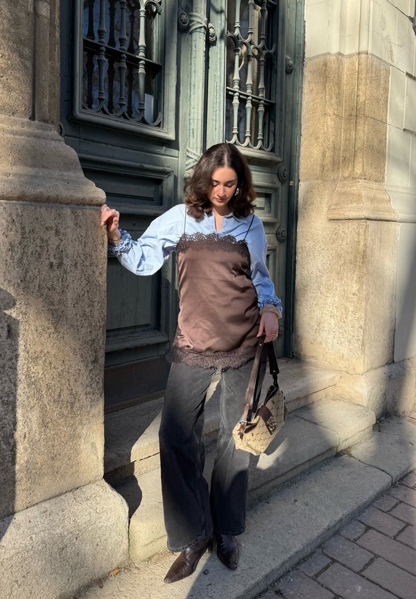 Joven con blusa de capas y camisola de satén marrón, vaqueros negros, botas marrones, sosteniendo un bolso con estampado, de pie junto a pilares de piedra y una puerta adornada.