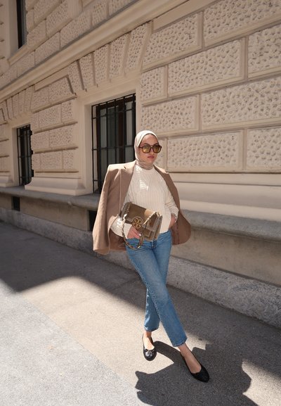 Blazer marrón, suéter de punto color crema, jeans azul claro, zapatos planos negros y un bolso con herrajes dorados. Las gafas de sol completan el look contra una pared texturizada.