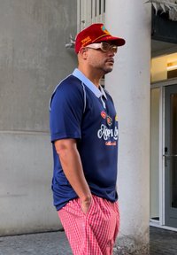 Polo shirt blu navy con dettagli bianchi e logo ricamato, abbinato a pantaloni a righe rosse e bianche. Cappellino rosso con logo e occhiali da sole.