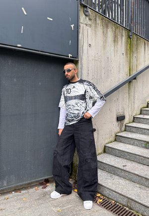 Hombre con gafas de sol, jersey deportivo extragrande y pantalones cargo negros anchos, de pie junto a una pared de hormigón y unas escaleras en un entorno urbano.