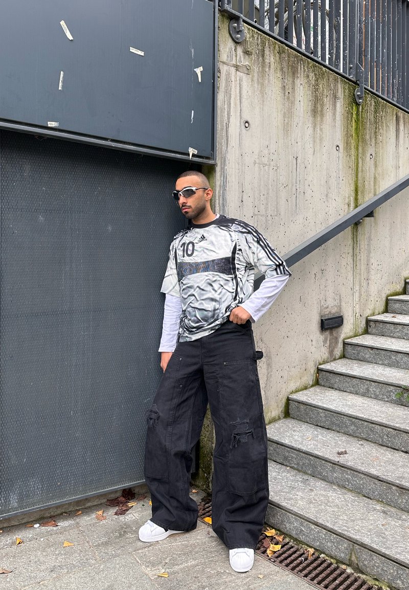 Άνδρας με γυαλιά ηλίου, oversized αθλητική φανέλα και φαρδιά μαύρα cargo παντελόνια, στέκεται δίπλα σε τσιμεντένιο τοίχο και σκάλες σε αστικό περιβάλλον.