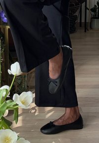 Bailarinas pretas com bico arredondado e um pequeno laço, feitas de couro liso, usadas por uma pessoa de pele escura, em pé ao lado de flores.
