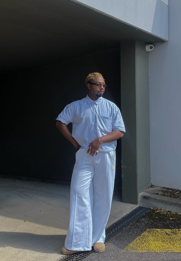 Camisa de manga corta azul clara con un bolsillo, combinada con pantalones anchos blancos. Los accesorios incluyen gafas de sol y un anillo. De pie contra una pared oscura.