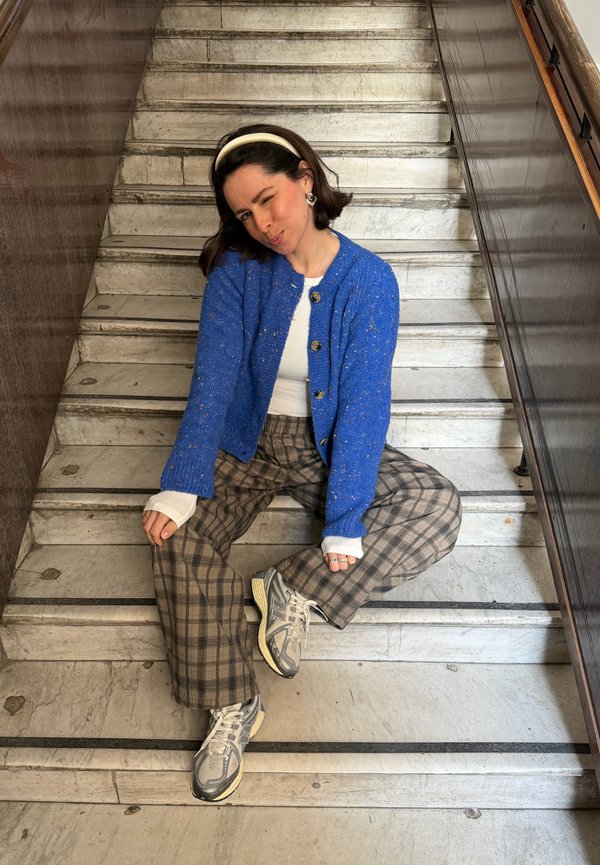 Jeune femme portant un cardigan bleu, un haut blanc, un pantalon à carreaux et des baskets, assise et faisant un clin d'œil sur des escaliers en marbre à l'intérieur.