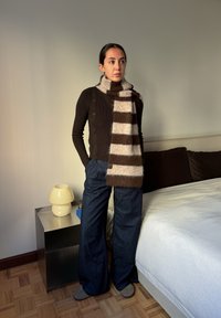 Hnědý žebrovaný svetr s knoflíky, pruhovaný béžový a hnědý šál, tmavě modré široké džíny a šedé kotníkové boty. Interiér pokoje s lampou.