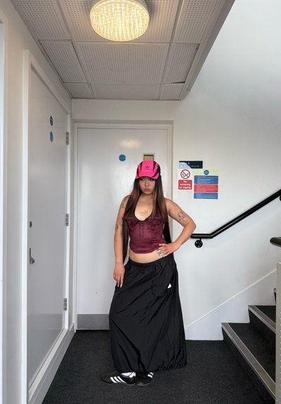 Top corto color marrón con un patrón texturizado, falda larga negra y sombrero rosa. Zapatos negros con rayas blancas completan el atuendo. Fondo de barandilla negra y puerta blanca.