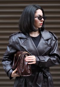 Brauner Trenchcoat aus Kunstleder mit großen Revers und Schulterklappen, kombiniert mit einer dunkelbraunen Handtasche mit goldfarbenen Akzenten.