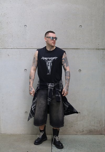 Camiseta sin mangas negra con estampado gráfico, combinada con pantalones cortos de mezclilla oscuros y holgados. Camisa de cuadros en blanco y negro atada a la cintura, botas negras.