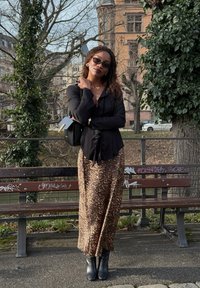 Femme portant un chemisier noir, une jupe à imprimé léopard, des bottes noires, des lunettes de soleil et un sac à épaule, debout devant un banc couvert de graffiti dans un parc urbain.