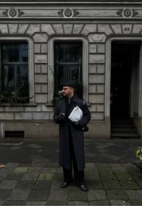 Άτομο που φοράει μαύρο μακρύ παλτό και καπέλο, κρατώντας μια εφημερίδα και καφέ, στέκεται σε μια πλακόστρωτη οδό μπροστά από ένα πέτρινο κτήριο με παράθυρα.