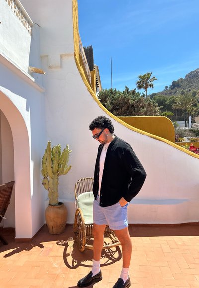 Cárdigan negro acanalado sobre una camisa blanca, pantalones cortos a rayas azules y zapatos negros. Cactus en una maceta de barro cercana; pared blanca y acentos amarillos.