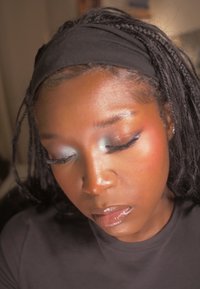 Gros plan sur un visage avec une peau lisse, un fard à paupières bleu métallisé, des lèvres brillantes, des cheveux tressés et un bandeau noir, portant un chemisier sombre.