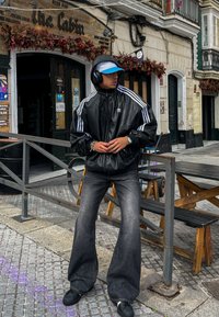 Svart lærjakke med glidelås og hvite striper, grå, vide jeans, svarte sko og hodetelefoner. Blåstripet caps som bæres bakfrem. Utendørs setting.