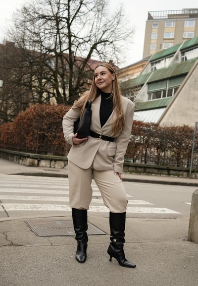 Mujer con traje beige y botas negras parada cerca del paso de peatones, sosteniendo un bolso negro, con árboles y edificios de fondo.