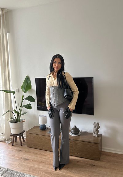 Mujer con cabello largo y negro, que viste blusa beige, corsé gris, pantalones grises, guantes negros y lleva un bolso negro, de pie en una sala de estar moderna.