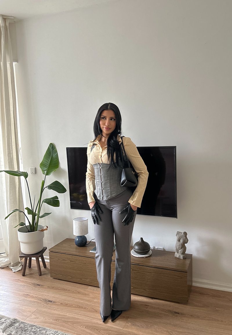 Femme aux longs cheveux noirs portant un chemisier beige, un corset gris, un pantalon gris, des gants noirs, et tenant un sac à main noir, debout dans un salon moderne.