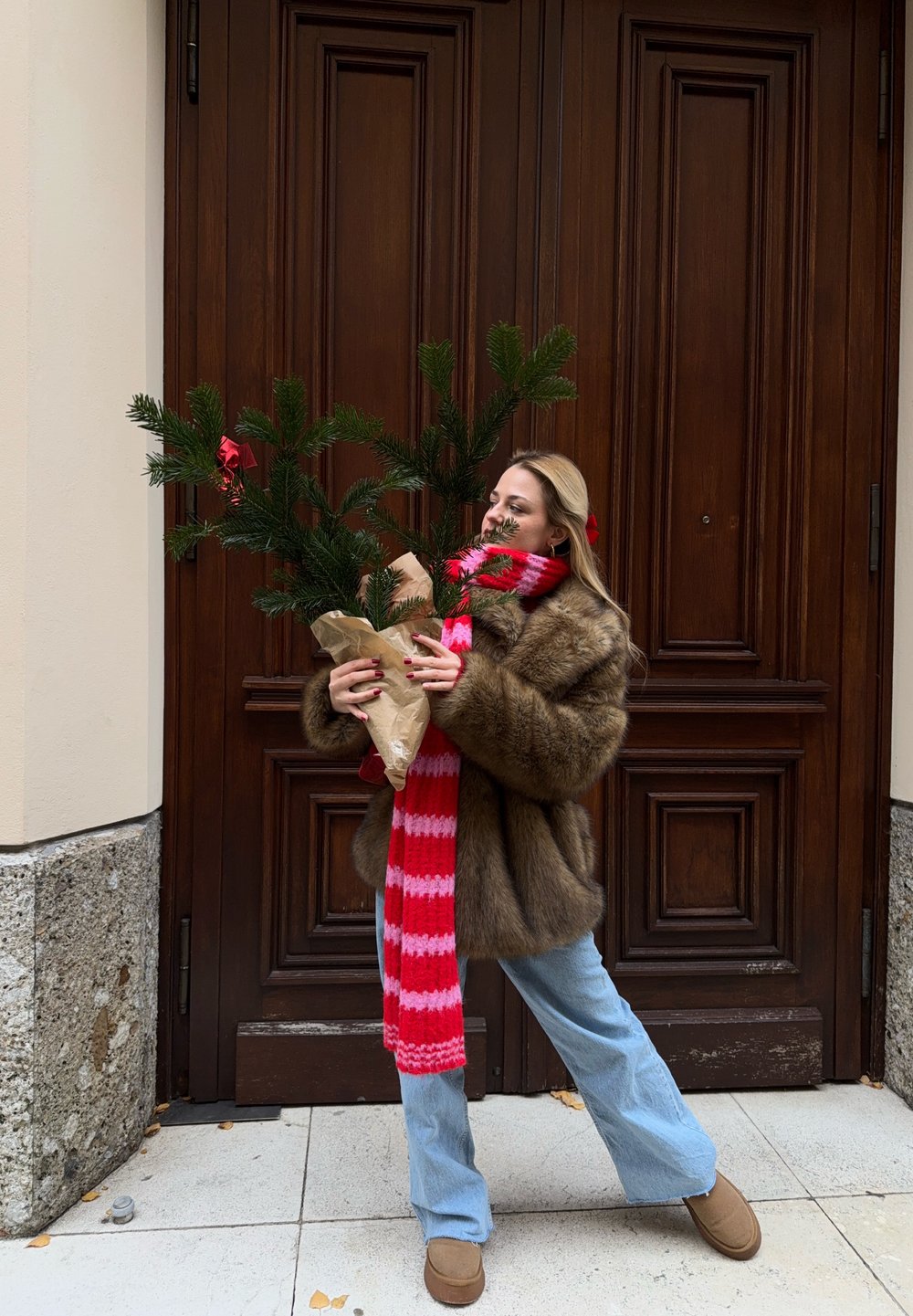 Faux fur coat, red and pink striped scarf, light blue flared jeans, holding a small potted pine tree wrapped in brown paper.