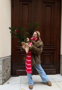 Faux fur coat, red and pink striped scarf, light blue flared jeans, holding a small potted pine tree wrapped in brown paper.