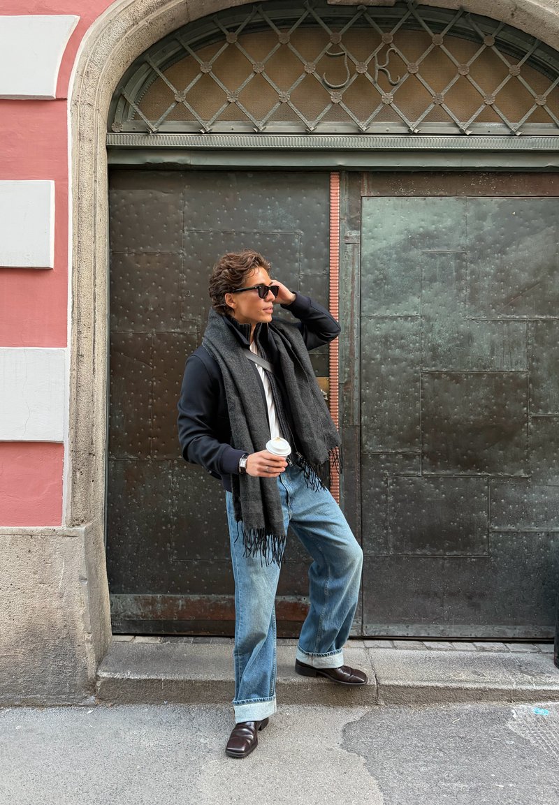 Pessoa segurando uma chávena de café, vestindo um lenço preto, casaco azul-marinho, camisa branca e jeans azul claro com as bainhas enroladas, parada perto de uma porta metálica texturizada.