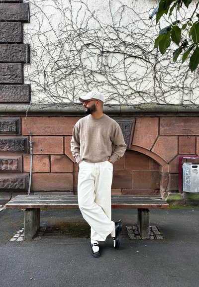 Suéter beige, pantalones de pierna ancha blancos y zapatos negros con acentos blancos. El fondo presenta una pared de piedra texturizada con enredaderas.
