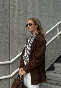 Brown suede oversized coat, striped shirt with a necktie, white trousers, and black sunglasses. Brown handbag. Concrete background with stairs.