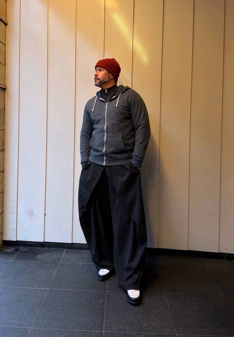 Un homme portant un bonnet rouge, un sweat à capuche gris zippé, un pantalon large à fines rayures et des chaussures blanches, debout contre un mur beige à panneaux.