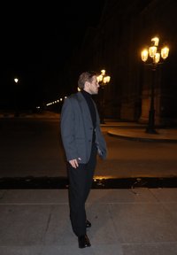 Grijze blazer over een zwarte coltrui, zwarte broek en schoenen. Avondsetting met straatlantaarns op de achtergrond. Strakke, gestructureerde lijnen.