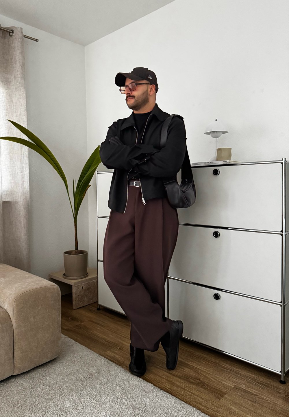 Μαύρο μπουφάν με φερμουάρ πάνω από μια μαύρη κορυφή, συνδυασμένο με καφέ φαρδιά παντελόνια και μαύρα παπούτσια. Μαύρο καπέλο και γυαλιά. Το φόντο περιλαμβάνει ένα φυτό και έπιπλα.