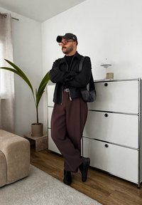 Μαύρο μπουφάν με φερμουάρ πάνω από μια μαύρη κορυφή, συνδυασμένο με καφέ φαρδιά παντελόνια και μαύρα παπούτσια. Μαύρο καπέλο και γυαλιά. Το φόντο περιλαμβάνει ένα φυτό και έπιπλα.