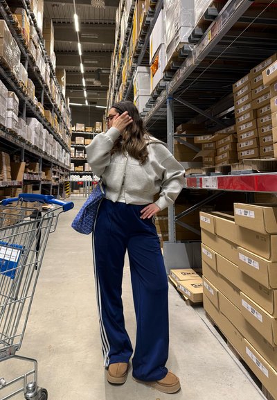Mujer con cabello largo y gafas está de pie en un pasillo de un almacén con estantes altos llenos de cajas, un carrito de la compra y una bolsa azul sobre su hombro.