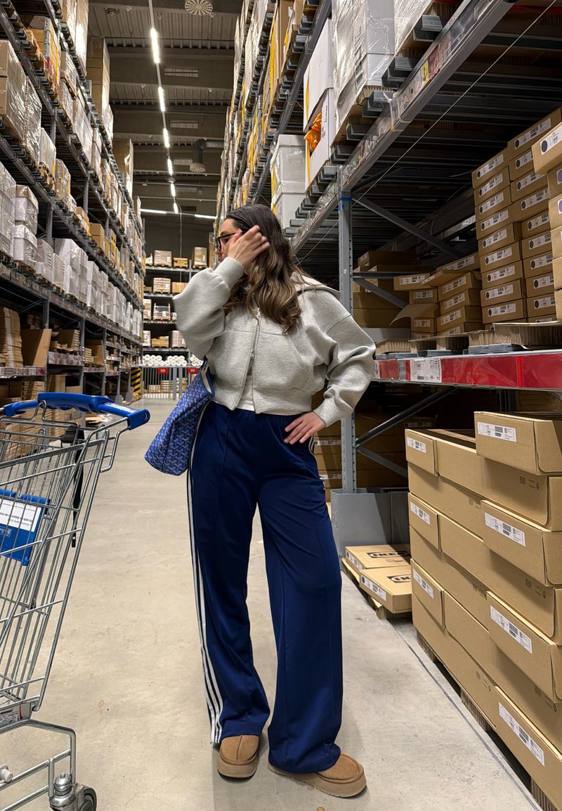 Femme aux longs cheveux et lunettes debout dans une allée d'entrepôt avec des étagères hautes remplies de cartons, un chariot de courses et un sac bleu sur l'épaule.