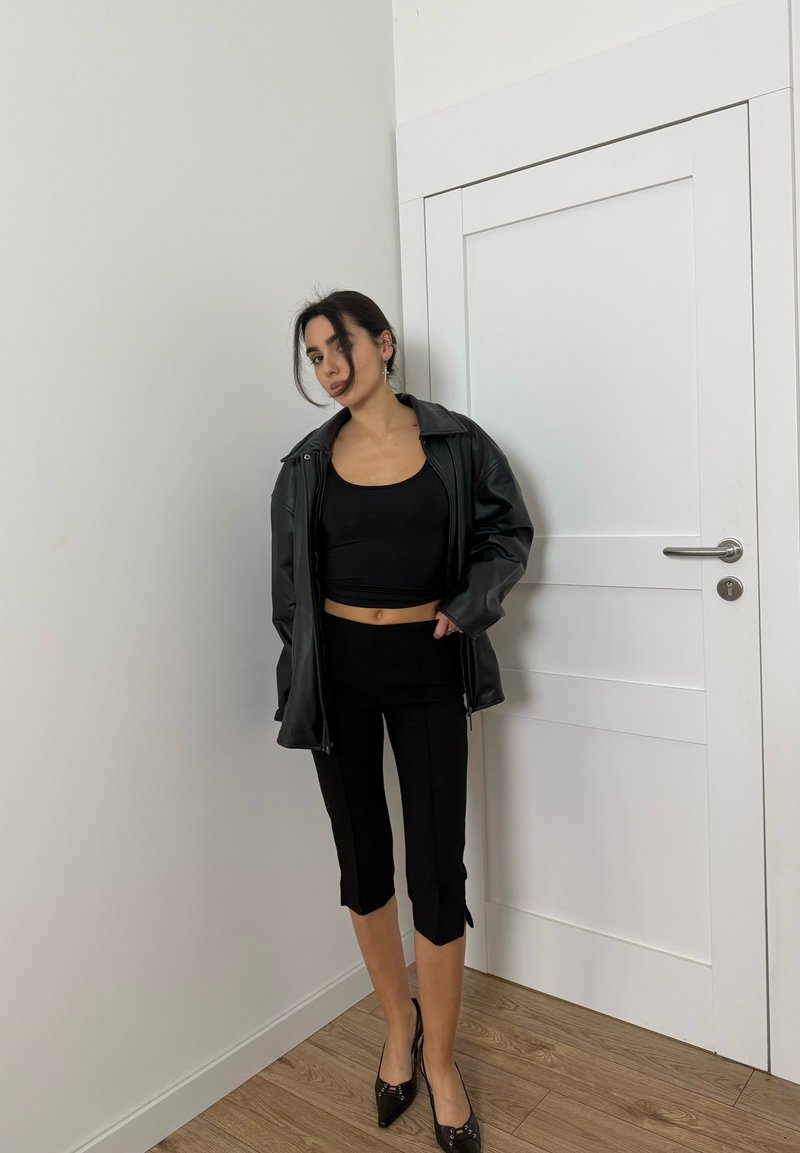 Femme debout à l'intérieur près d'une porte blanche, portant un crop top noir, une veste en cuir noire, un pantalon capri noir et des escarpins pointus noirs.